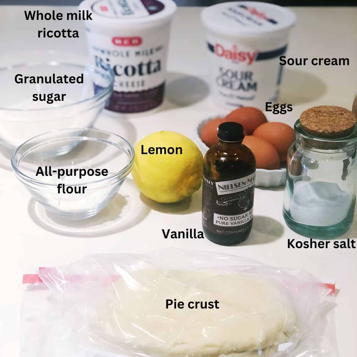 Ricotta pie ingredients on the counter including: pie crust, ricotta cheese, lemon, sour cream, vanilla , sugar, flour, eggs and salt.