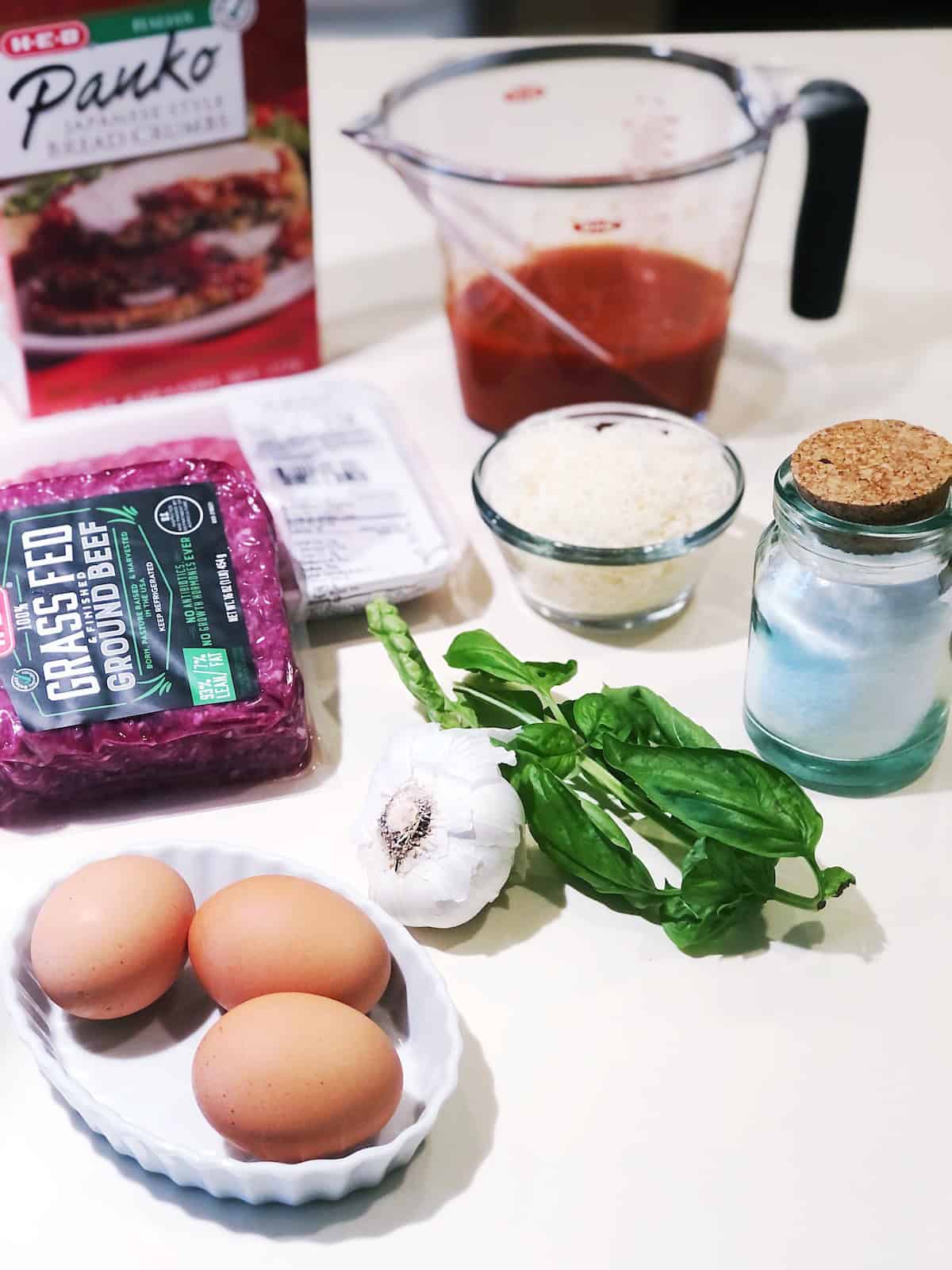 Italian meatloaf ingredients on the counter including, eggs, basil, garlic, breadcrumbs, ground beef, Parmesan, salt and marinara.
